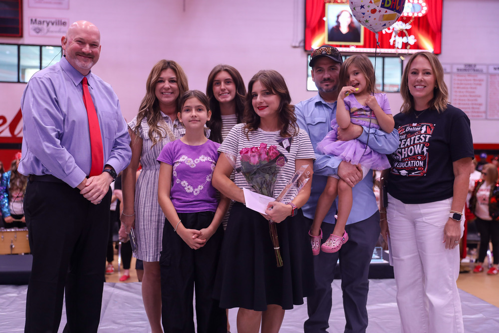 A woman holding a bouquet of pink roses stands at the center of a stage inside a gymnasium alongside her family and two school administrators. The man on the far left wears a light blue dress shirt with a red tie, while the woman on the far right wears a black "The Greatest Show in Education" t-shirt and white pants. In the background, a large screen displays a picture of the honoree.
