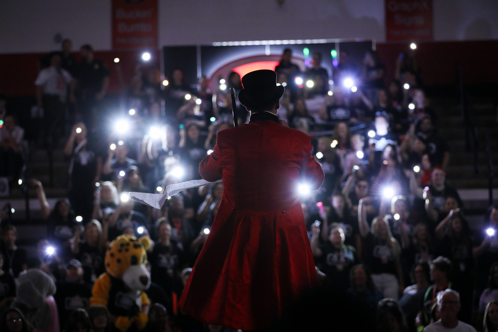A performer seen from behind wearing a red ringmaster's coat and top hat stands on stage facing an indoor arena crowd. The audience in the bleachers holds up phone flashlights, creating dozens of bright spots of light across the dim room. Below to the left, a yellow jaguar mascot costume is visible among the spectators.