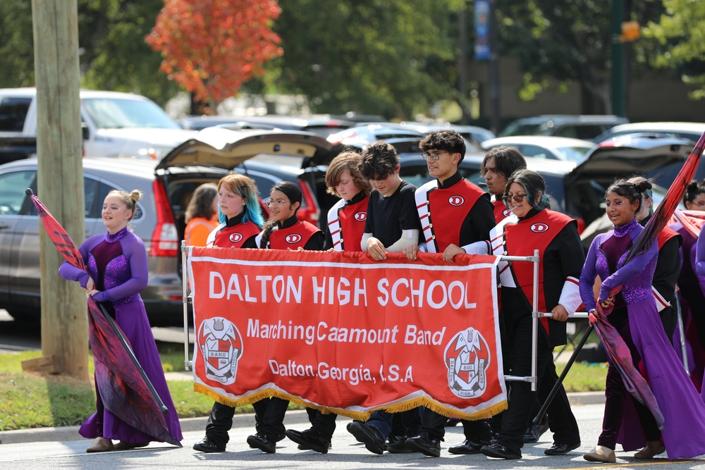 A group of high school students marches down an outdoor street lined with parked vehicles. In the center, students dressed in red, black, and white band uniforms hold up a large red banner reading "DALTON HIGH SCHOOL Marching Catamount Band Dalton, Georgia, U.S.A." Color guard members wearing purple dresses march alongside them holding flags on poles.