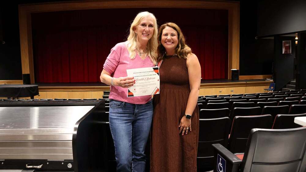 Two educators stand together in a school auditorium, smiling while one holds a certificate recognizing an achievement in education. Rows of auditorium seats and a stage with a red curtain are visible in the background. The image captures a moment of recognition and celebration, highlighting excellence in teaching and the districtās commitment to honoring outstanding educators.
