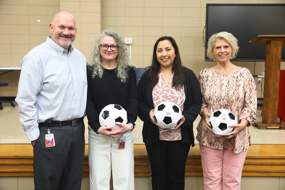 Four adults stand together smiling in a school meeting or gym space. Three women hold black-and-white soccer balls labeled āDalton Game Ball,ā while a man on the far left wears a light button-down shirt and a Dalton Public Schools ID badge. The women are dressed in business-casual attire, and the group stands in front of beige block walls, a stage area, and a large display screen.