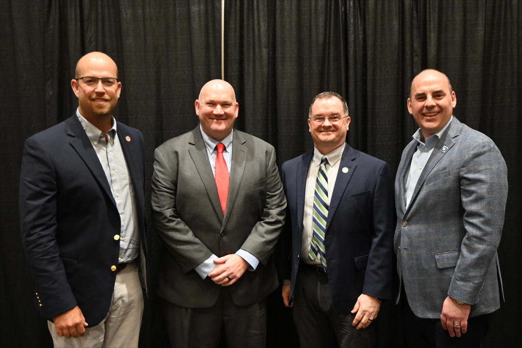 Four men in suits and blazers stand side by side in front of a black curtain, smiling for the camera.