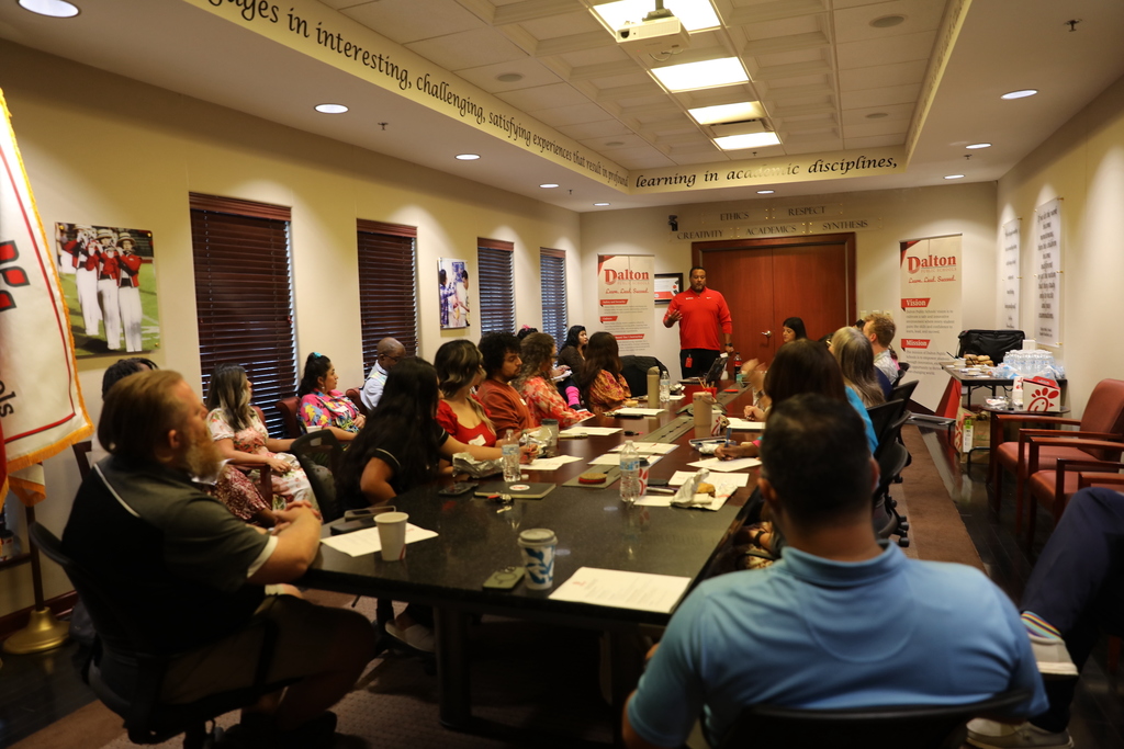 A large group of parents, staff, and community members sit around a long conference table at Dalton Public Schools during a meeting. A speaker in a red shirt addresses the group at the front of the room, while attendees listen and take notes. Food, drinks, and papers are on the table.