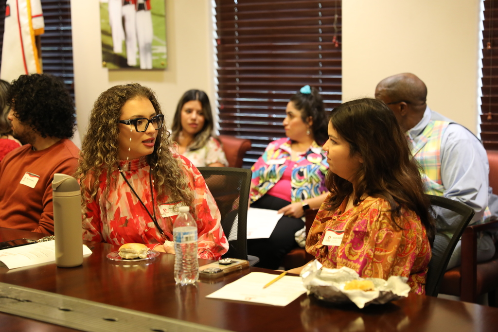 Participants at the Dalton Public Schools meeting engage in discussion around the conference table. A woman in a red patterned blouse speaks with another attendee, while others listen and take notes.