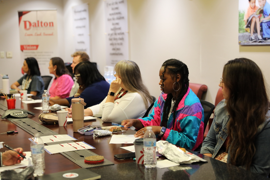 Several attendees sit on the other side of the conference table, reviewing papers and eating breakfast during the Dalton Public Schools meeting. A banner with the district’s mission and vision is displayed in the background.