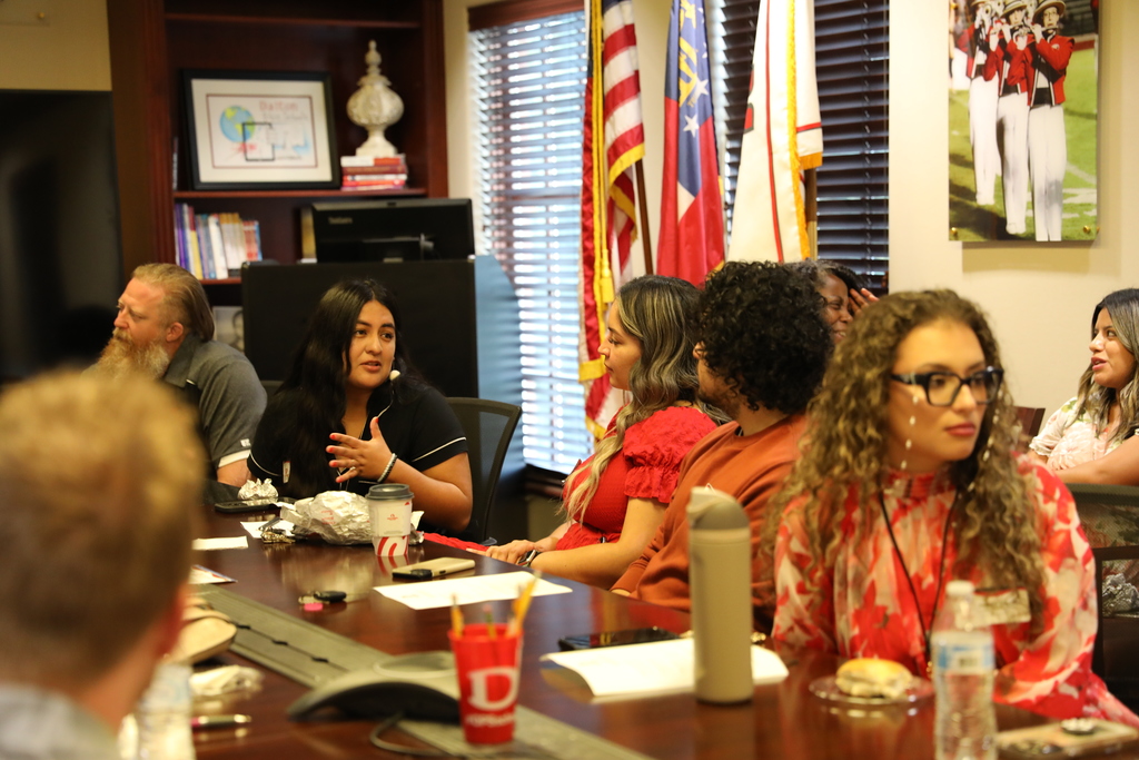 A woman speaks while gesturing during the Dalton Public Schools meeting, seated among a group of attendees. Flags and framed photos are displayed in the background, and others listen as the discussion continues.