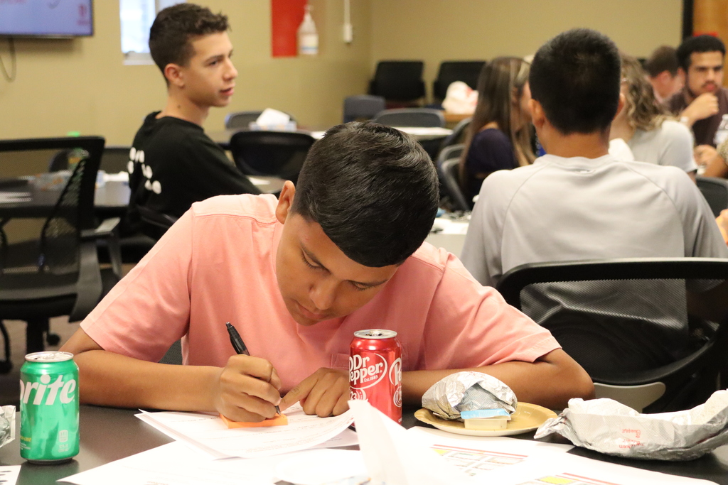 A student writes notes on paper during the meeting, with soda cans and lunch items on the table. Other students are visible in the background.