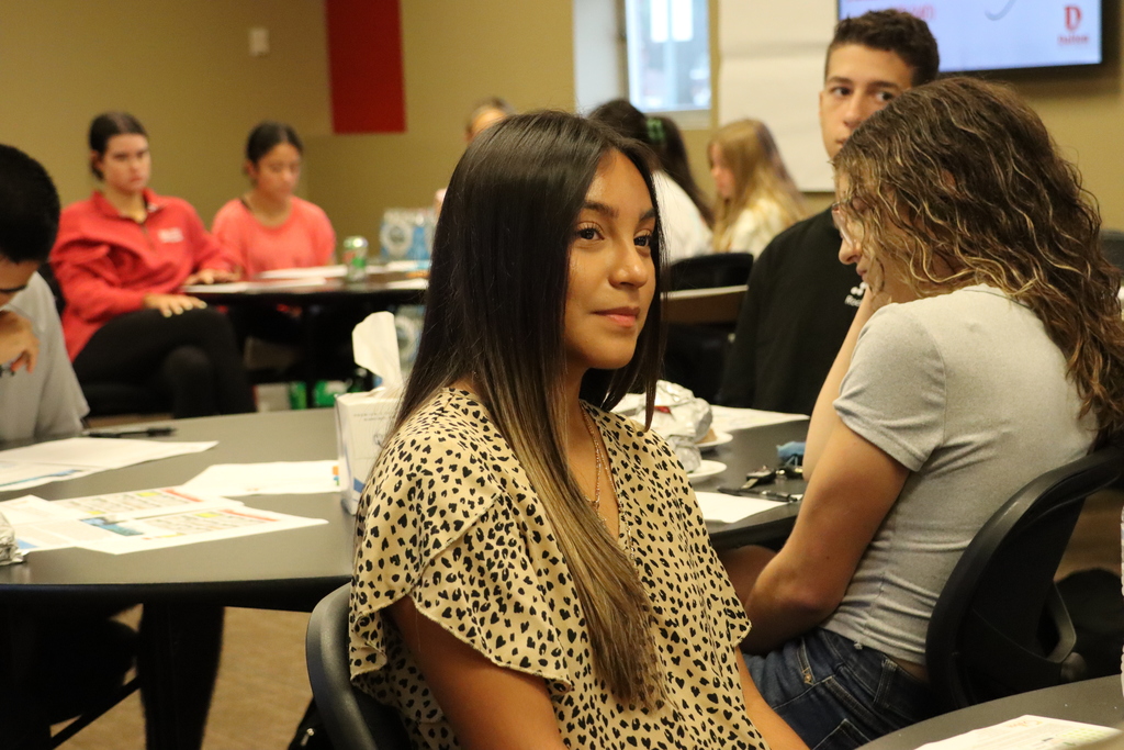 A student in a patterned shirt smiles at the camera while other Student Advisory Council members sit at round tables behind her.