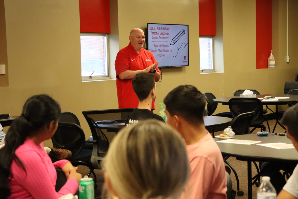 Superintendent Dr. Craft speaks to the Student Advisory Council while standing in front of a presentation screen about Dalton Public Schools’ personal electronic device procedure. Students listen attentively around tables.