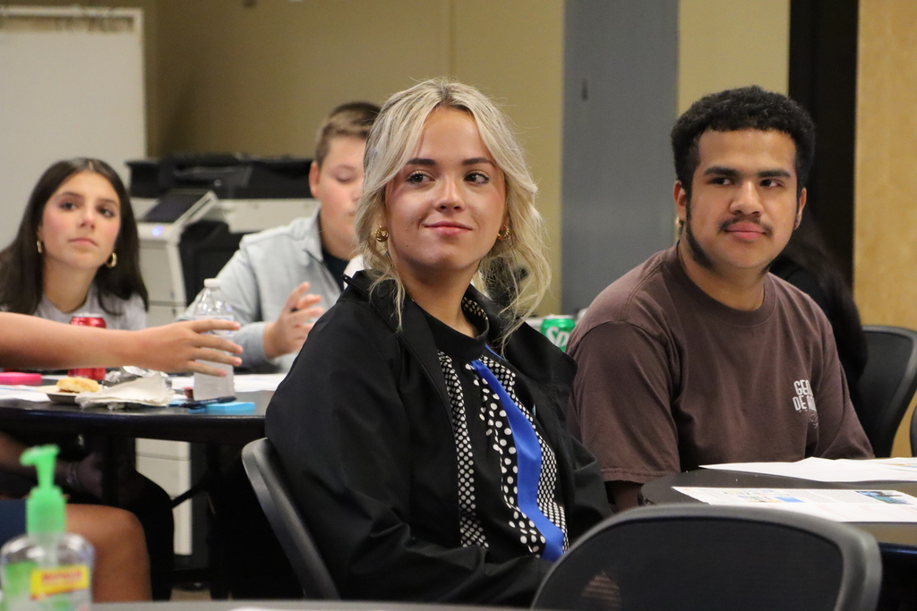 Two students smile and listen during the Student Advisory Council meeting, with other students visible in the background.