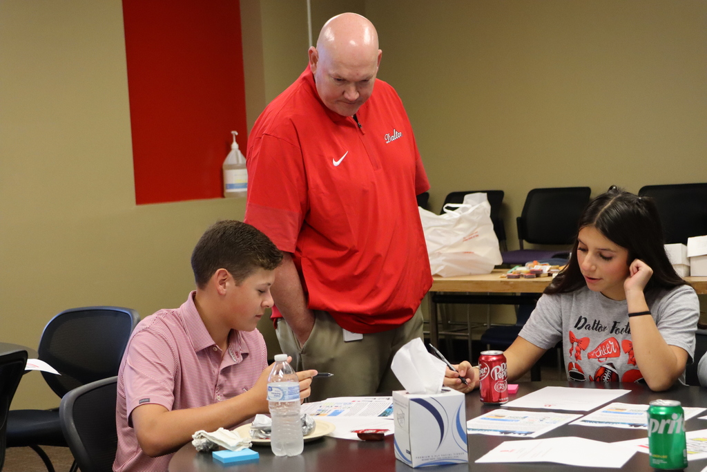 Superintendent Dr. Craft leans over a table to talk with two students who are working on documents during the meeting.