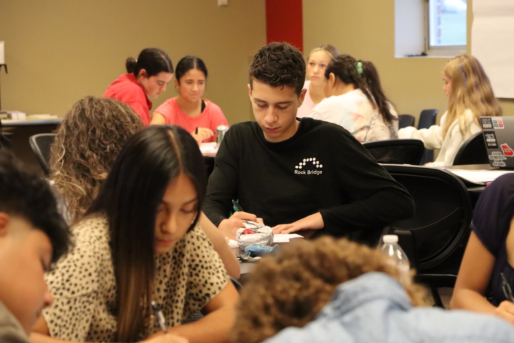 A student focuses on writing while other Student Advisory Council members work and talk in the background.
