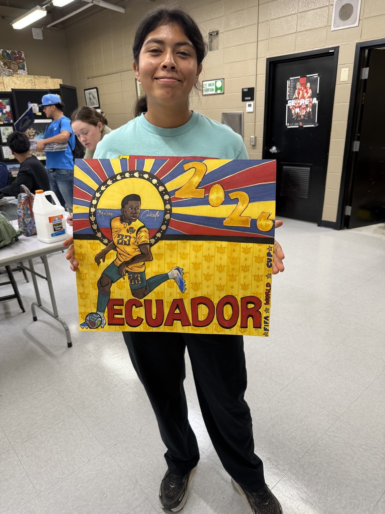 A student stands in an art classroom holding a bright yellow, blue, and red artwork representing Ecuador for the 2026 World Cup. She is smiling softly and wearing a teal T-shirt and black pants. The artwork depicts an Ecuadorian soccer player in action with the number 23 on the jersey, set against radiating sunburst stripes in the colors of the Ecuadorian flag. The large text “2026” appears at the top right, and “ECUADOR” is written boldly along the bottom. Other students are visible working in the background at tables.
