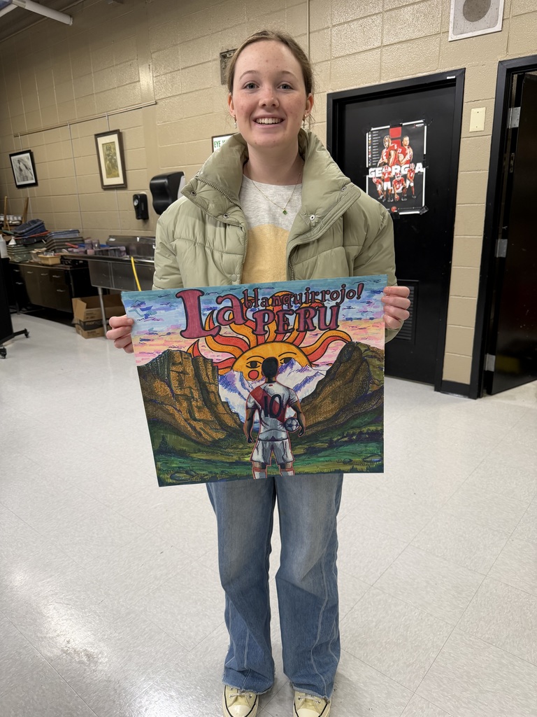 A student stands in a classroom holding a colorful artwork representing Peru for the 2026 World Cup. She is smiling and wearing a light green puffer jacket over a beige shirt with blue jeans. The artwork features a soccer player wearing a Peru jersey with the number 10, standing in a valley framed by mountains. A bright sun with a face rises behind the peaks, and stylized red and pink lettering at the top reads “La blanquirroja! PERU.” The classroom background includes tan brick walls, framed art on the walls, stacked supplies, and stainless-steel sinks.