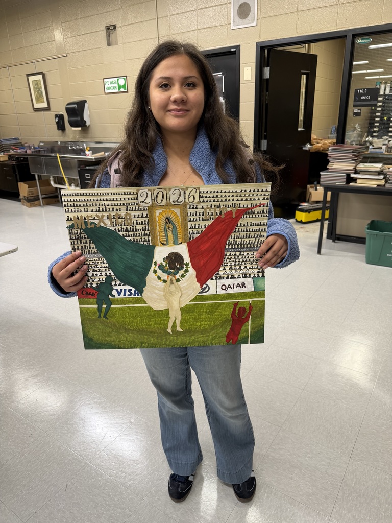 A student stands in a classroom holding an artwork representing Mexico City for the 2026 World Cup. She is smiling and wearing a fuzzy blue zip-up jacket and light blue jeans. The artwork shows a soccer player holding a large Mexican flag on a field, with stadium stands packed with spectators behind them. At the center of the art piece is an image of the Virgin of Guadalupe framed by a golden archway. The large text “2026” appears above the scene. The classroom behind her includes shelves of art supplies, books, and materials.