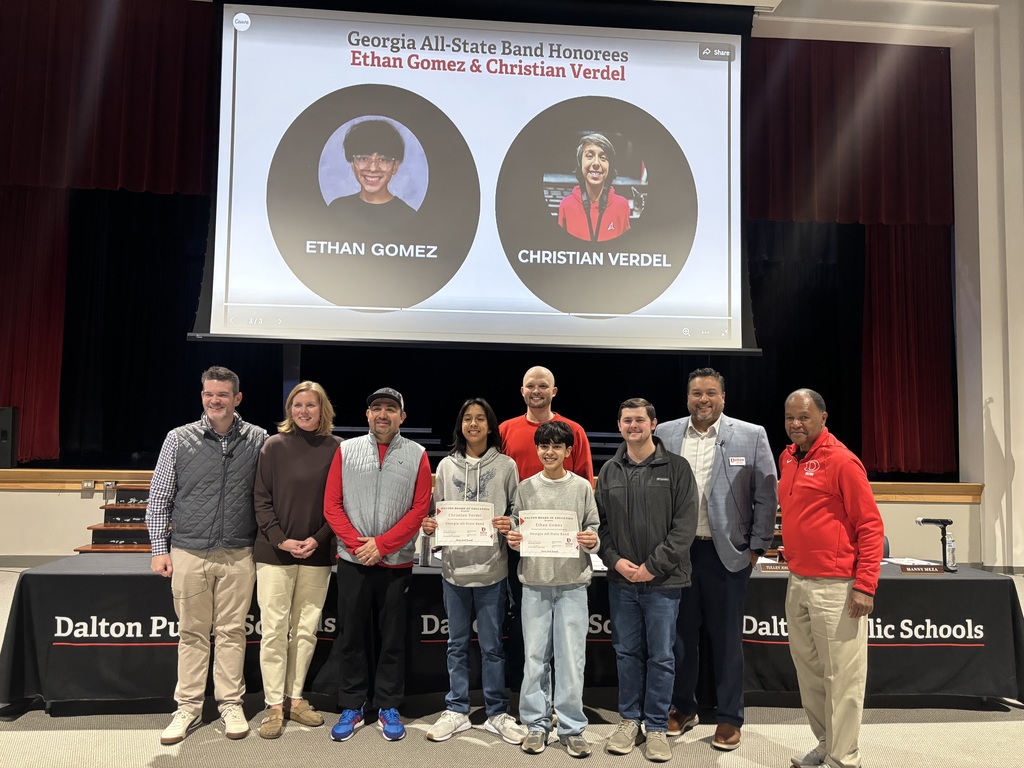 Two Dalton Public Schools students stand at the center of a group of district leaders and staff during a Board of Education meeting, holding certificates recognizing their selection to the Georgia All-State Band. A large screen behind them displays their names and photos, labeled “Georgia All-State Band Honorees: Ethan Gomez & Christian Verdel.” The group is posed on a stage in a school auditorium with Dalton Public Schools signage visible on the table in front of them.
