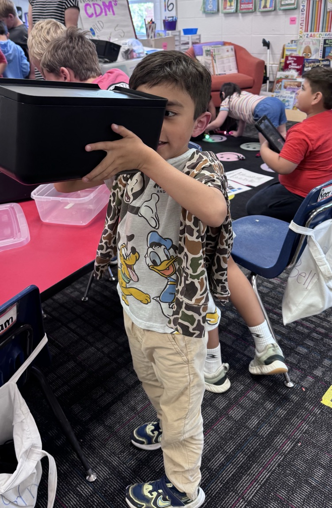 A young student in a classroom holds up a black viewing device to his eyes, smiling as he explores an activity.