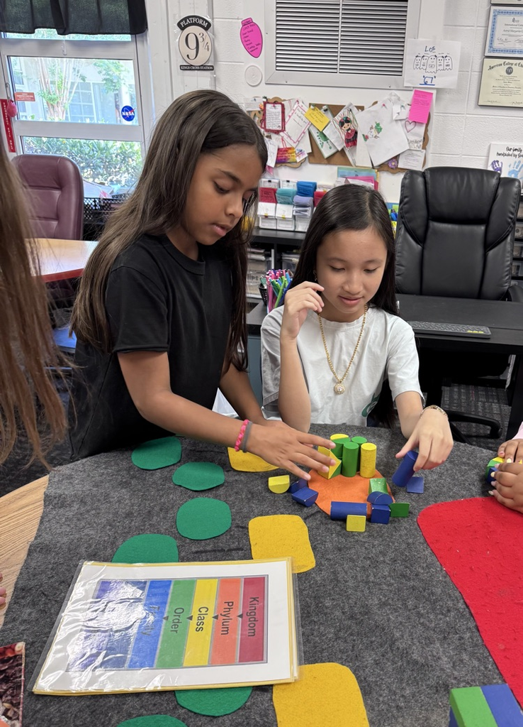 Two students work together at a classroom table, using colorful geometric blocks to build and explore. One student carefully places pieces while the other observes and thinks through the activity.