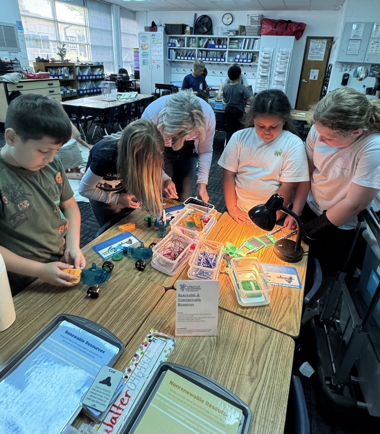 Students gather around a classroom table working on a hands-on STEM activity with a teacher. They explore renewable and nonrenewable resources using small building components, wires, and a light source, while reading informational cards.