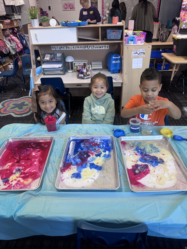 Three young students sit at a classroom table, smiling and engaged in a colorful hands-on activity. In front of them are trays filled with vibrant mixtures of red, blue, and yellow, creating swirling patterns as they experiment.