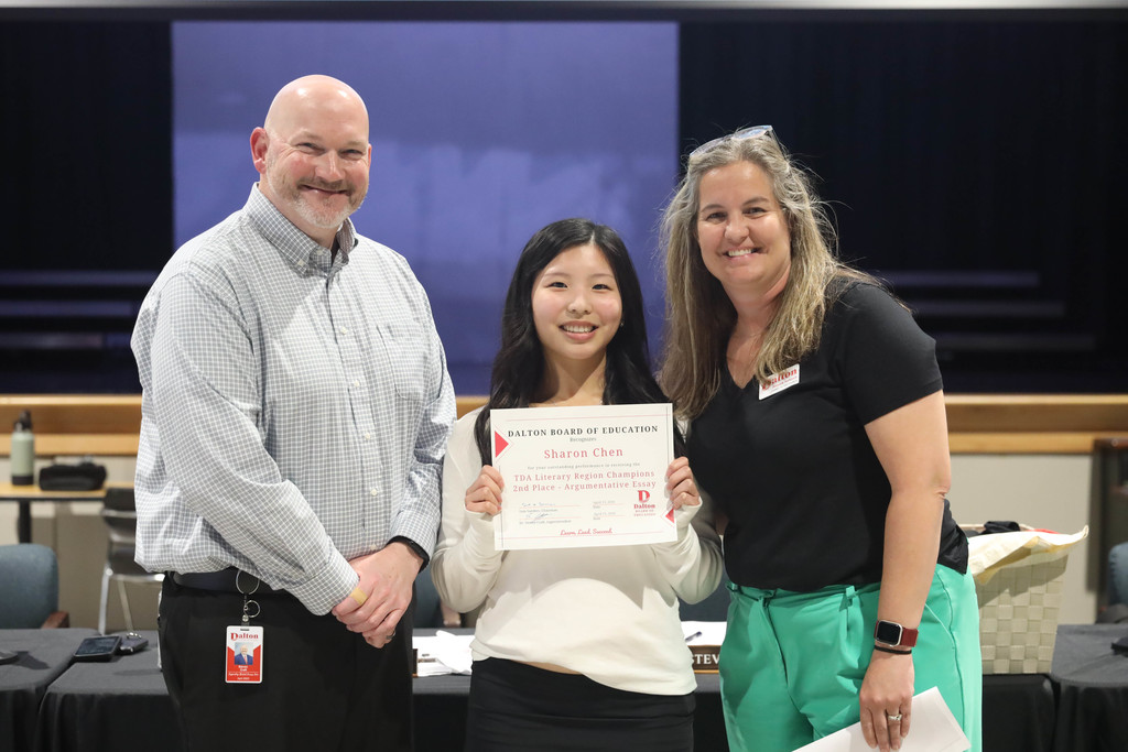 A student stands between two adults at a Dalton Public Schools boardroom, smiling while holding a certificate that recognizes her achievement. The adults on either side also smile, and a meeting table with school materials and signage is visible in the background.