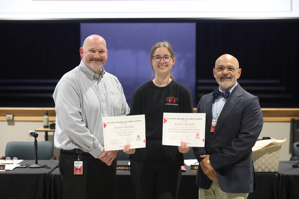 A student stands between two school officials in a Dalton Public Schools boardroom, holding two certificates recognizing her achievements. All three are smiling at the camera, with a meeting table, microphones, and nameplates visible in the background.