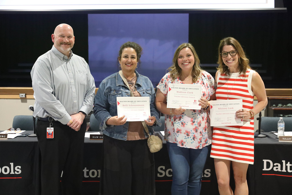 Four adults stand together at a Dalton Public Schools board meeting, smiling and holding certificates of recognition. Three women hold awards while a man stands beside them, with a boardroom setting and Dalton Schools signage visible in the background.