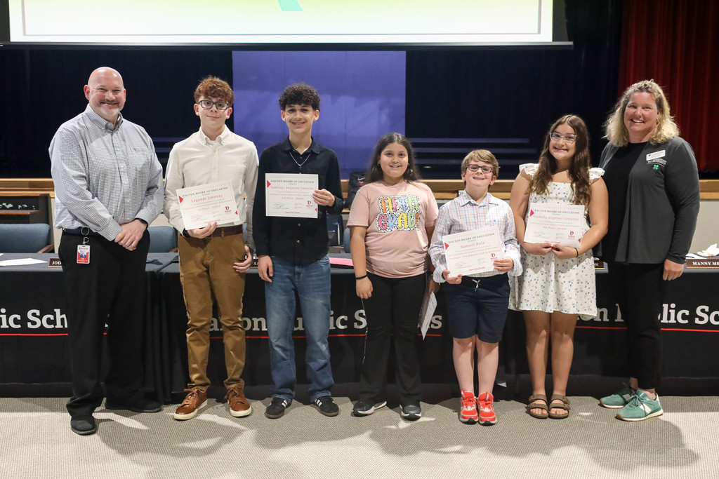 A group of students and two adults stand together at a Dalton Public Schools boardroom, smiling and holding certificates of recognition. The students display their awards while standing in a line, with a meeting table and Dalton Public Schools signage visible in the background.