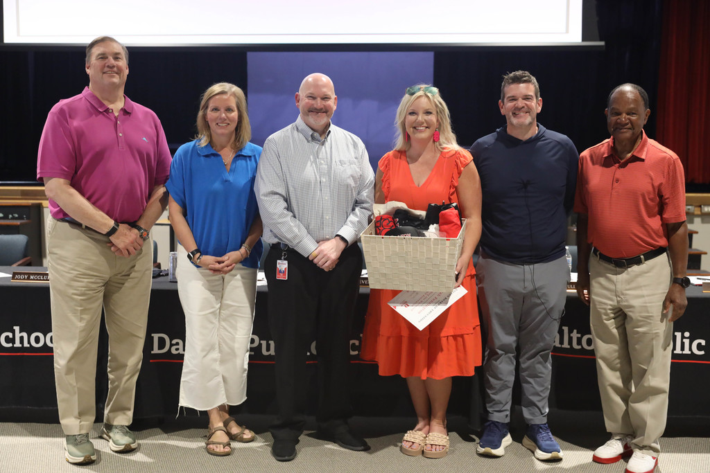 Six adults stand together at a Dalton Public Schools boardroom, smiling at the camera. One woman holds a gift basket and papers, suggesting a recognition or appreciation presentation, with Dalton Public Schools signage visible behind them.