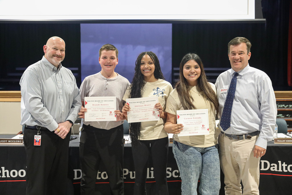 Three students stand with two school officials in a Dalton Public Schools boardroom, each student holding a certificate of recognition. All five are smiling and facing the camera, with a meeting table, microphones, and nameplates visible in the background.