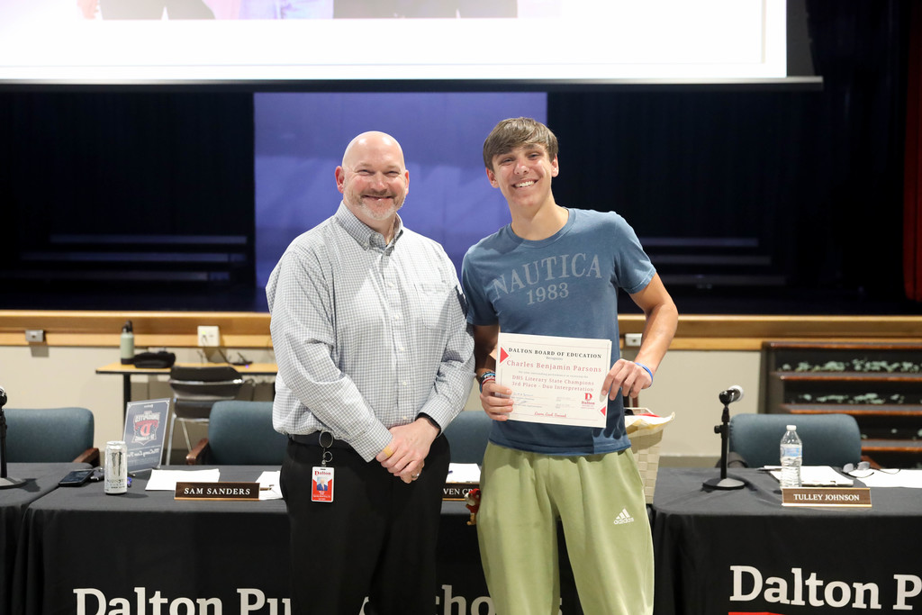 A smiling teenage student stands beside a school official at a Dalton Public Schools boardroom, holding a certificate of recognition. Both face the camera, with a meeting table, microphones, and nameplates visible behind them.