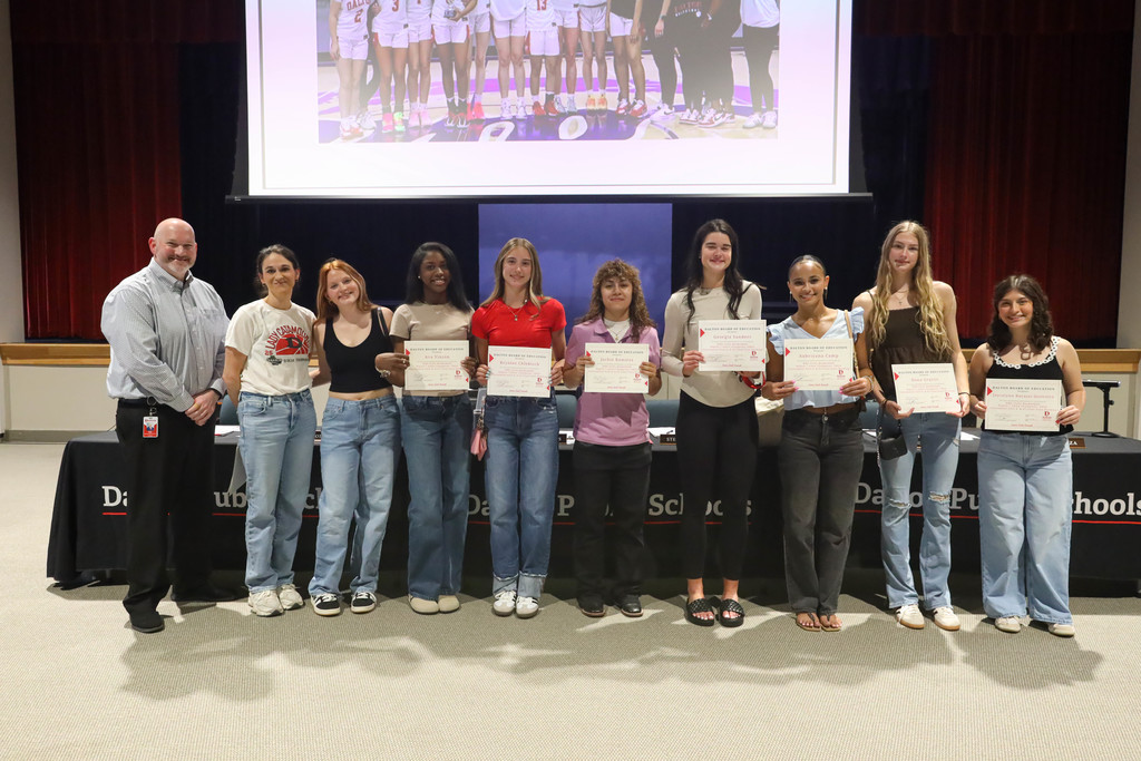 A group of students stand in a line with a school official in a Dalton Public Schools boardroom, each student holding a certificate of recognition. They are smiling at the camera, with a meeting table, microphones, and a projection screen visible behind them.