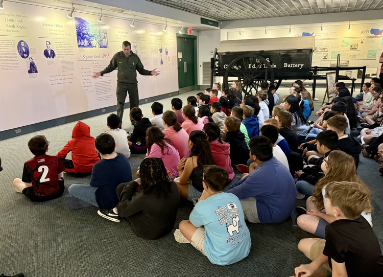 Students sit on the floor listening to a park ranger giving a history presentation inside a museum, with a Civil War wagon exhibit behind him.