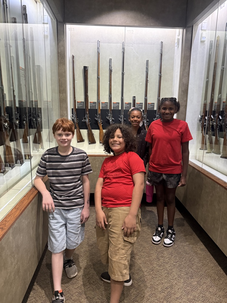 Four students smile and pose in a museum hallway with historic rifles displayed in glass cases behind them.