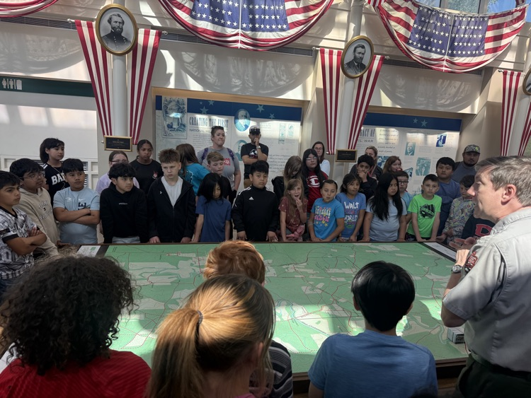 Students gather around a large map display as a park ranger explains a Civil War site inside a museum with American flags overhead.
