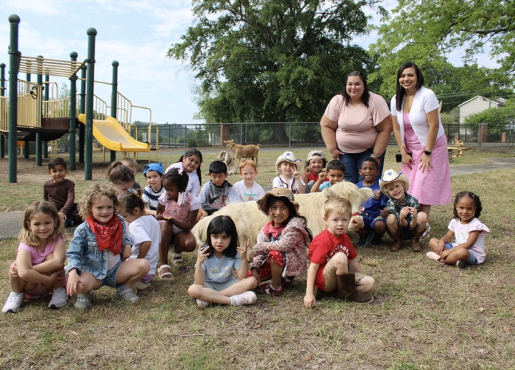A group picture of students sitting on the ground around the cow and 2 teachers standing  