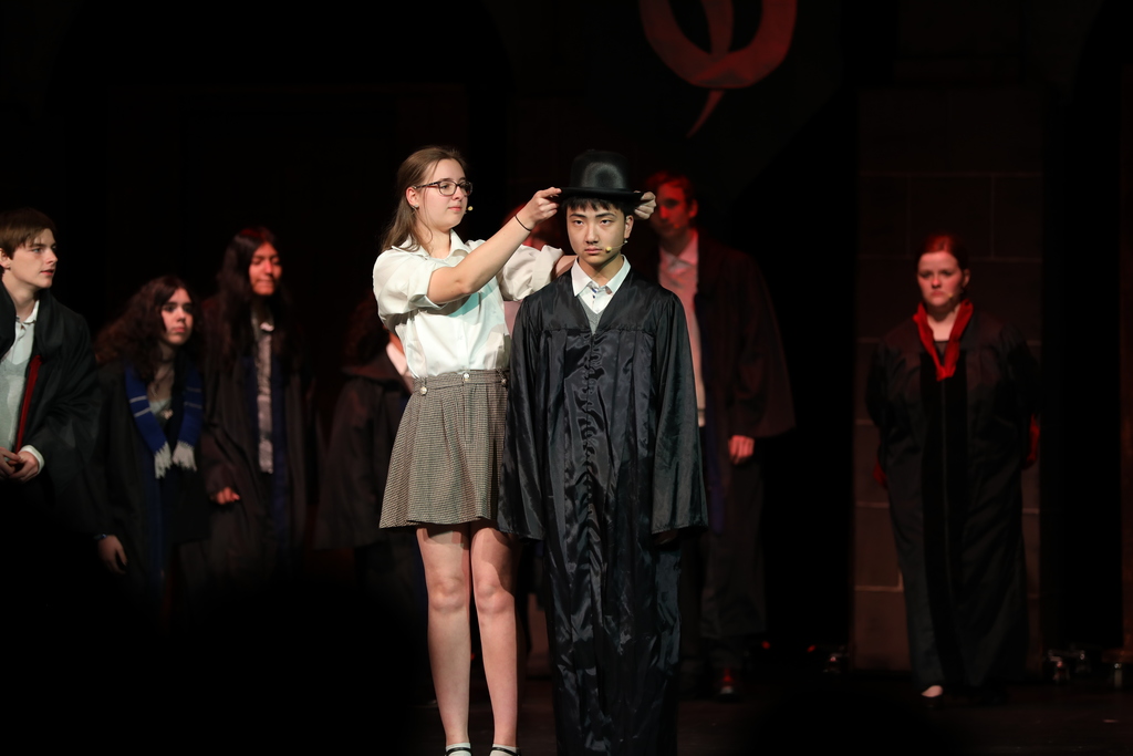 A student places a black hat on another student dressed in a long black robe during a stage performance. Several other students in costume stand in the background, watching. The scene is lit with warm stage lighting, suggesting a ceremonial or dramatic moment in the play.
