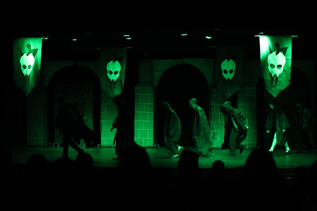 A group of students in hooded cloaks perform on a dimly lit stage under green lighting. They move in a line with hunched postures, creating a dramatic, eerie scene. The backdrop features archways and hanging banners with skull-like symbols, enhancing the dark, theatrical atmosphere.