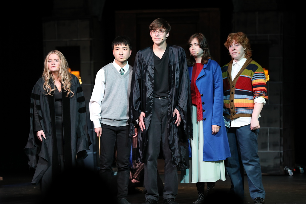 Five students stand on a stage during a theatrical performance, wearing costumes and microphones. The central student wears a black robe, while others are dressed in varied outfits including a sweater vest, a blue coat, and a colorful striped cardigan. They appear serious and focused under stage lighting with a dark set in the background.
