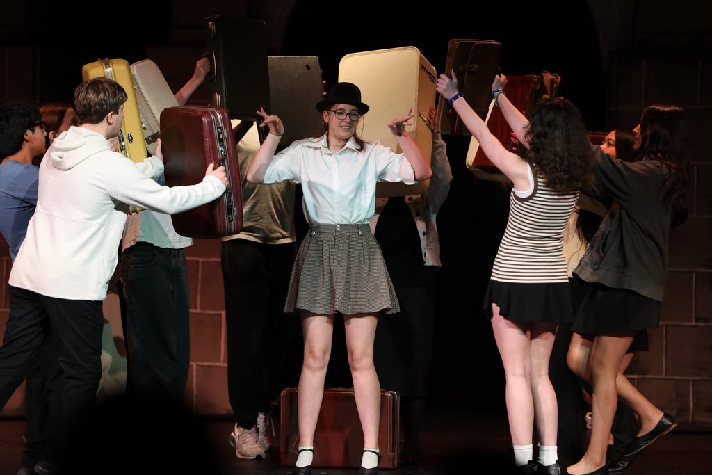 Students perform a lively scene on stage, with one student standing center wearing a white shirt, skirt, and bowler hat, posing confidently. Around them, other students hold up various suitcases, creating a dynamic, choreographed moment. The stage lighting highlights the performers against a dark backdrop.