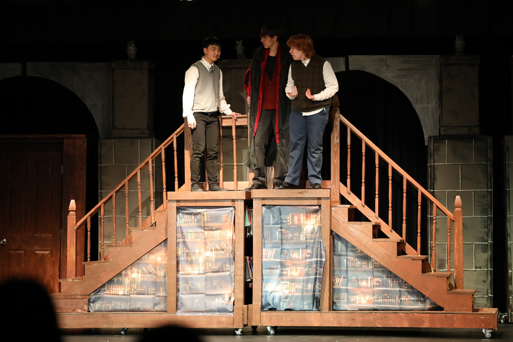 Three students stand on a raised stage set with wooden stairs and bookshelves, performing a scene under warm lighting. One student in a long dark coat stands in the center, flanked by two others in sweater vests, as they appear to be in conversation or reacting to something. The background features arched set pieces, creating a dramatic theatrical setting.