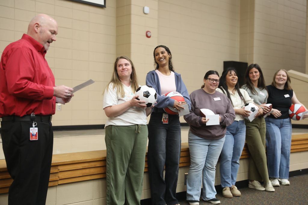 A group photo inside a school gym or meeting space featuring six young women and one man standing together and smiling at the camera. Several students are holding sports balls, including soccer balls, footballs, and red-and-white “Dalton Game Ball” basketballs. The man on the far left is wearing a red button-down shirt and black pants with a Dalton Public Schools ID badge. The RBTs wear casual clothing and name tags, standing in front of a beige block wall and wooden risers.
