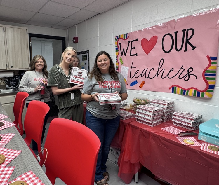 Three teachers holding their pizzas in front of the “We love our teachers” sign in the teachers lounge 