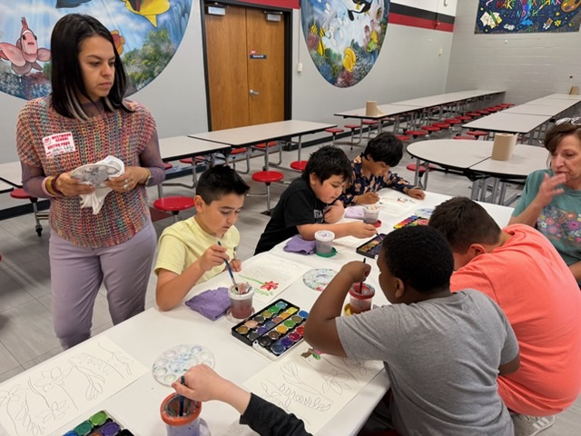 Students sitting in the cafeteria painting flowers while Mayelli Meza walks around to check their paintings