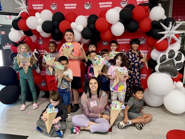 A pictures of the group of students holding their painted bouquets of flowers with Mrs. Meza in front of the Westwood backdrop 