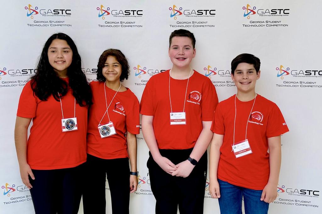 Four students stand smiling in front of a Georgia Student Technology Competition (GASTC) backdrop. They are all wearing matching bright red “Innovators Club Technology Team” T-shirts and name badges on lanyards. The group includes two girls and two boys standing side by side, dressed casually in dark pants and jeans. The white backdrop behind them is covered with repeating GASTC logos and the words “Georgia Student Technology Competition.”