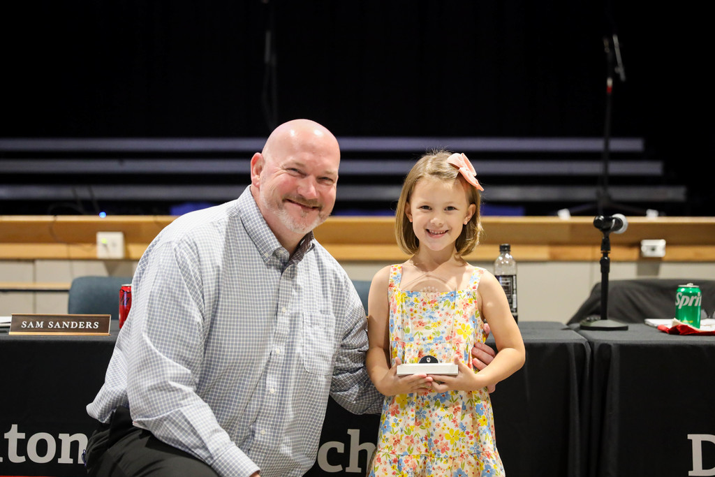 A smiling young girl in a colorful floral dress with a pink bow in her hair stands beside a smiling man in a light checkered button-down shirt at a school recognition event. The girl holds a small award or plaque while the man kneels slightly beside her with one arm around her back. Behind them are tables with microphones, drinks, and nameplates, including one reading “Sam Sanders,” set in an auditorium or board meeting space.