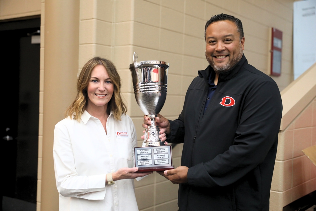 Two school staff members smile while holding a large silver trophy together inside a school building. The woman on the left wears a white button-down shirt with a Dalton logo, and the man on the right wears a black jacket with a red āDā emblem. The trophy sits on a wooden base with engraved plaques listing school names and years. Beige block walls and a staircase are visible in the background.