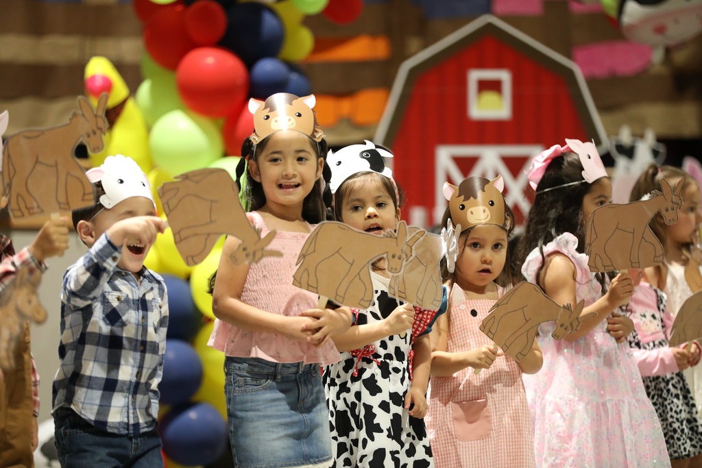 A group of young children stand on a decorated stage during a school performance, wearing handmade farm-animal headbands and holding paper animal cutouts on sticks. The children are dressed in colorful outfits, including plaid shirts, denim, dresses, and cow-print clothing. Behind them are bright balloons and a large red barn backdrop, creating a cheerful farm-themed scene.