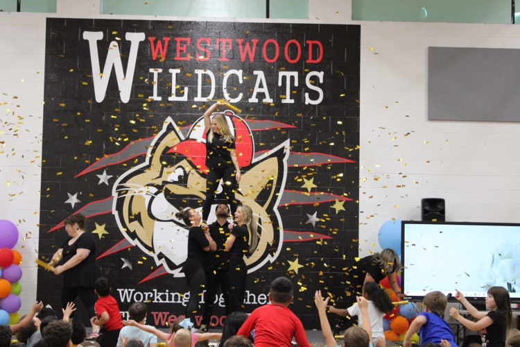 Teachers perform during the talent show as students cheer and gold confetti fills the air in front of the Westwood Wildcats mural.